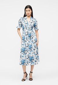 Γυναίκα που φοράει λευκό midi φόρεμα με μπλε floral σχέδιο, φουσκωτά μανίκια, V-λαιμόκοψη και μαύρα ψηλοτάκουνα σανδάλια, που στέκεται μπροστά σε απλό φόντο.