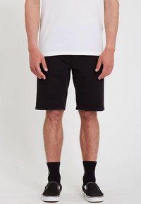 Black denim shorts with a straight cut, slight fraying at the hem, worn with black socks and slip-on sneakers. White t-shirt on top.