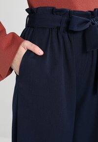 Marinblå högmidjade byxor med ett brett bälte som har en rosett. Tyget är slätt med en lätt lyster; har sidofickor och veck vid midjan.