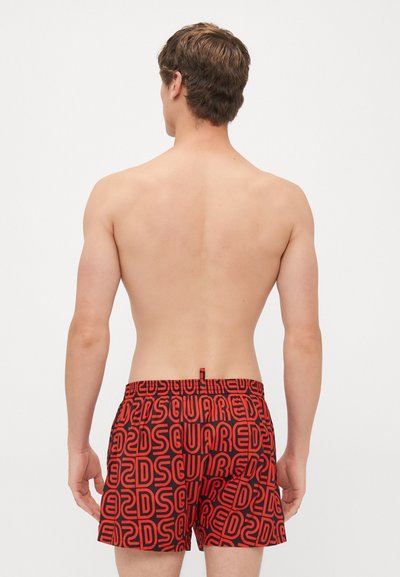 Homme au dos nu portant un short noir et rouge à motif avec le texte "DSQUARED2" en superposition, se tenant devant un fond blanc uni.