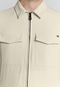 Beige ritsjack met een kraaghalslijn, twee borstzakken en zwarte ritsdetails. Gladde textuur en modern ontwerp.