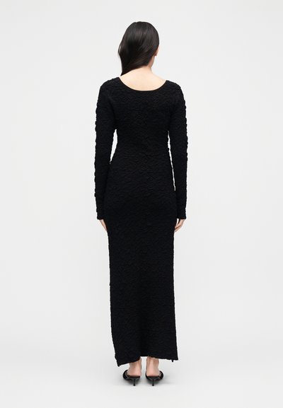 Frau mit langen dunklen Haaren, die ein strukturiertes, schwarzes, langärmliges, eng anliegendes Maxikleid und schwarze Kitten-Heel-Sandalen trägt, mit dem Rücken zur Kamera vor weißem Hintergrund.