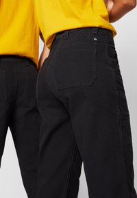 Pantaloni in denim neri con vita alta, dotati di due tasche posteriori e cuciture a contrasto. Il completo è abbinato a una maglietta gialla.