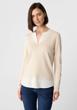 Femme aux cheveux bruns courts portant un pull beige à col en V texturé sur une blouse blanche et un jean bleu, debout devant un fond uni.