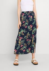 Floral maxi skirt in navy, featuring pink and green patterns, an elastic waistband, and a side slit. Worn with white sandals.