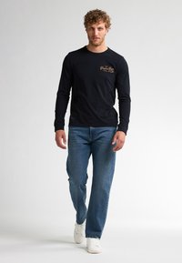 Zwarte longsleeve shirt met gouden logodetail, gecombineerd met lichtblauwe rechte jeans en witte sneakers, gedragen door een staande mannelijke model.