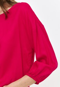 Fuksjowa bluzka o luźnym kroju, z okrągłym dekoltem i marszczonymi rękawami do łokcia. Gładki materiał o delikatnym połysku.
