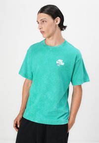 T-shirt de algodão verde com um corte descontraído, mangas curtas, gola redonda e um logótipo branco da Nike Air no lado esquerdo do peito. Acabamento suave e texturizado.