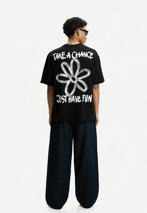 Sort oversized t-shirt med hvid tekst: "TAG EN CHANCE" og "HAV BARE DET SJOVT" ved siden af et stort hvidt blomster grafik. Båret med vide denim bukser.