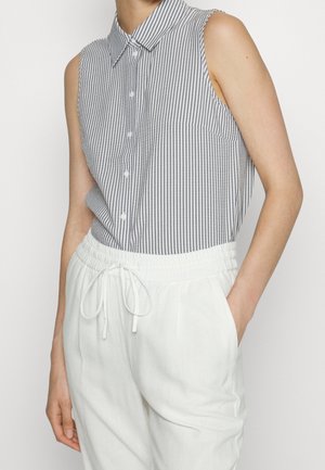 Chemise sans manches rayée verticalement gris et blanc, rentrée dans un pantalon blanc à cordon avec taille élastique, une main dans la poche.