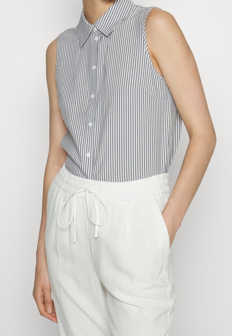 Chemise sans manches rayée verticalement gris et blanc, rentrée dans un pantalon blanc à cordon avec taille élastique, une main dans la poche.