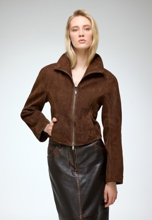 Brown suede jacket with a high collar, zip front, structured shape, and wide sleeves, paired with a fitted leather skirt.