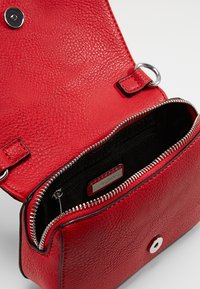 Sac bandoulière en cuir rouge texturé avec fermeture éclair argentée et bouton-pression magnétique, montrant le compartiment principal ouvert et la poche intérieure zippée portant l'étiquette ALDO.