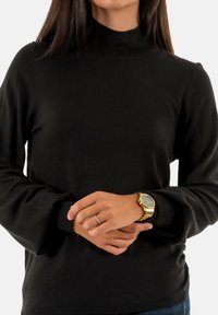 Suéter negro de cuello alto hecho de una tela suave, con puños y cuello acanalados. Manos juntas, llevando un reloj de oro.