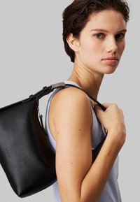 Sac à main en cuir noir au design minimaliste, doté d'une fermeture éclair et d'une bandoulière ajustable. Texture lisse avec un détail de marque subtil.