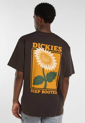 Persona con camiseta marrón oscuro con un gran gráfico de girasol y texto "DICKIES DEEP ROOTED" en la espalda sobre un fondo liso.