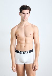 Weiße Boxershorts mit einem schwarzen "DIESEL" Logo-Bund, die eine glatte Textur und eine taillierte Passform aufweisen. Das Model steht in neutraler Pose.