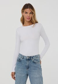 Top bianco a maniche lunghe realizzato in tessuto leggero, con scollo rotondo. Abbinato a jeans in denim blu a vita alta con un finish sbiadito.