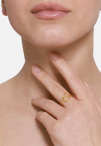 Goldring mit verwobenem Design, ausgestattet mit einer glatten, polierten Oberfläche. Die Hand des Models berührt sanft ihren Hals.