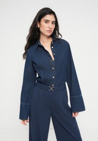 Τζιν πουκάμισο με κουμπιά, γιακά και μακριά μανίκια, με χρυσά κουμπιά, σε συνδυασμό με σκούρο μπλε παντελόνι ψηλόμεσο με λεπτομέρεια ρουμπινιού στη ζώνη.