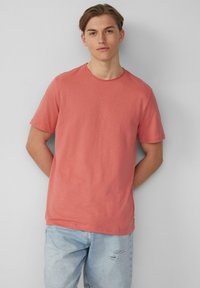 Korte mouwen, ronde hals t-shirt in koraalkleur, gemaakt van zachte stof. Heeft een relaxte pasvorm en een gladde textuur, gecombineerd met lichtblauwe distressed jeans.