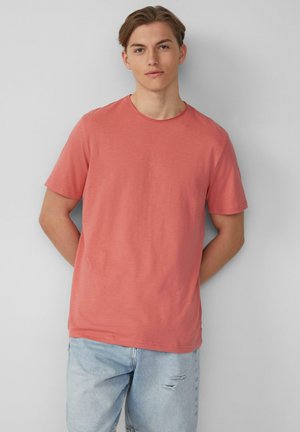 Korte mouwen, ronde hals t-shirt in koraalkleur, gemaakt van zachte stof. Heeft een relaxte pasvorm en een gladde textuur, gecombineerd met lichtblauwe distressed jeans.