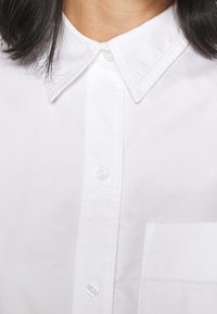 Ingrandimento del colletto di una camicia bianca con cuciture visibili e bottoni superiori, indossata da una persona con capelli neri di lunghezza alle spalle.