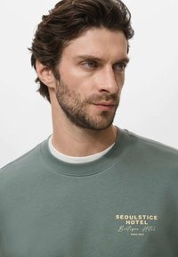 Green cotton sweatshirt with a crew neck, featuring "SEOULSTICE HOTEL Boutique Hotel" text in gold print on the chest. Smooth texture.