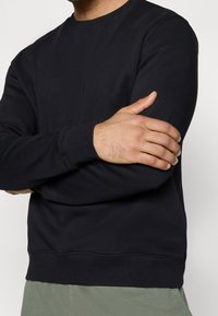 Sweat-shirt noir à col rond en tissu doux, avec manches longues et poignets côtelés. Le bas est pourvu d'un ourlet côtelé à texture lisse.