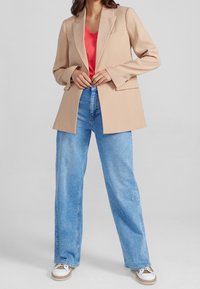 Béžový oversize blejzr s dlouhými rukávy, jednou kapsou, spárovaný se světle modrými širokými kalhotami a bílými teniskami s hnědými akcenty.