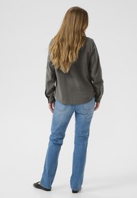 Camicia grigia a maniche lunghe con orlo arrotondato, abbinata a jeans dritti azzurri. Il modello ha capelli lunghi e mossi e indossa ballerine nere.