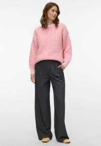 Femme portant un pull rose à motifs, un pantalon noir ample et des chaussures beige, debout avec une main dans la poche sur un fond blanc.