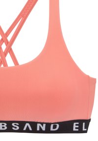 Bra sportivo in corallo con spalline incrociate, texture liscia e fascia nera a contrasto con branding bianco attorno al fondo.