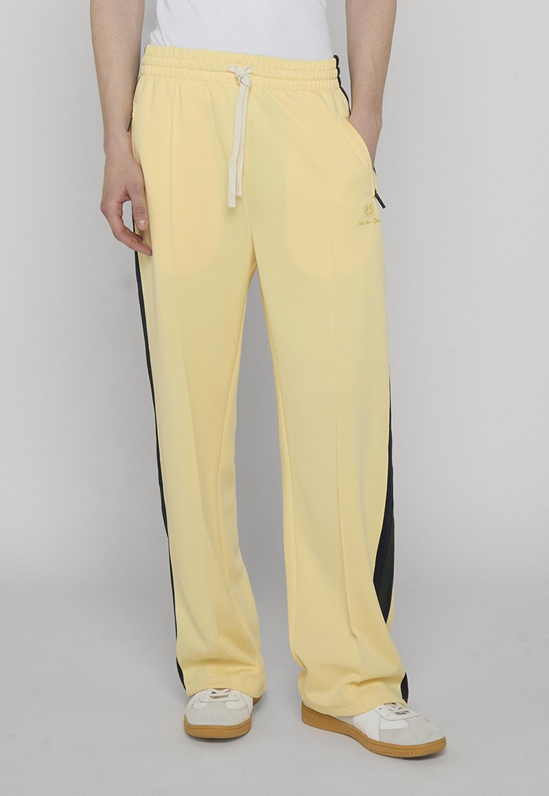 Alakeha seljas kahvatukollased jooksupüksid musta küljeribaga, valge nööriga ja valged tennised kummipõhjadega hallil põrandal.