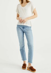 Top bianco a portafoglio con tessuto testurizzato, maniche corte e vita arricciata; jeans azzurri a gamba dritta; zoccoli marroni con accenti dorati.