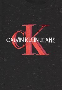 Černé tričko s výrazným červeným a růžovým vyšívaným logem "CK" a textem "CALVIN KLEIN JEANS", na pozadí jemně strukturované tkaniny.