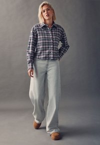 Plaid button-up shirt in purple, gray, and white; light gray wide-leg jeans; tan footwear. Model standing against a gray background.