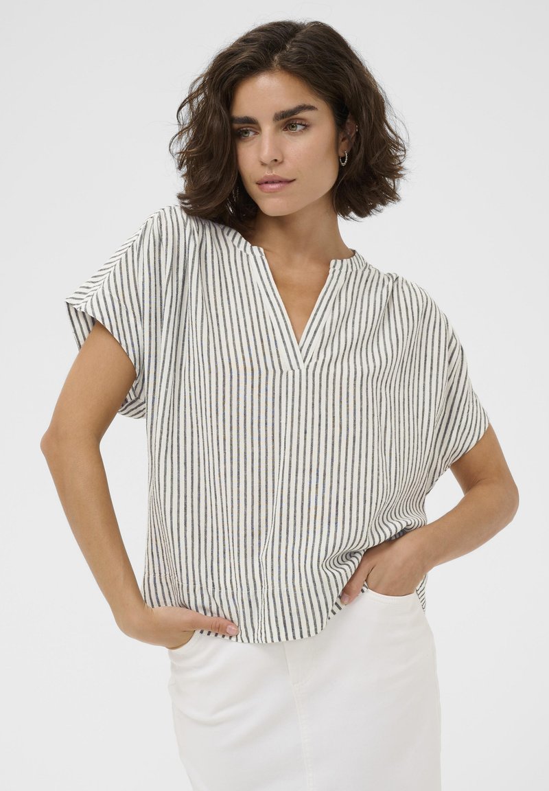 Femme aux cheveux courts et foncés, portant un haut à manches courtes rayé verticalement noir et blanc et un pantalon blanc, les mains dans les poches, sur un fond uni.