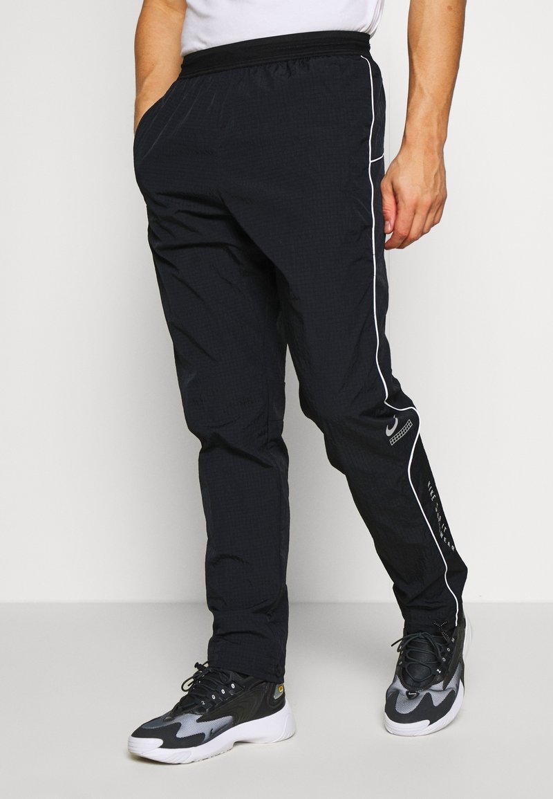 Man i svarta Nike sportbyxor med vit kantning och svarta och vita träningsskor står mot en enfärgad bakgrund.