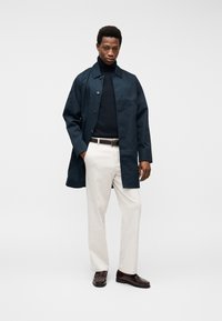 Άνδρας με μπλε σκούρο παλτό πάνω από μαύρο ζιβάγκο, λευκό παντελόνι με καφέ ζώνη και σκούρα καφέ μοκασίνια, στέκεται μπροστά σε μονόχρωμο φόντο.