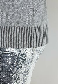 Maglione grigio a maglia con orlo marrone a costine, abbinato a pantaloni di paillettes argentate riflettenti, che mostrano una finitura lucida e testurizzata.
