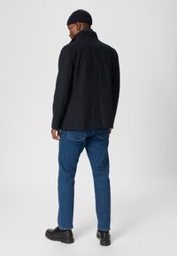 Marinblå ullblandad jacka med hög krage, i kombination med blå denimjeans och svarta läderskor. Jackan har en skräddarsydd passform och ett enkelt sprund.