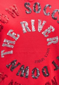 Red cotton t-shirt featuring circular design made of sequined letters in silver and black, with text detailing "SOCCER" and "RIVER."