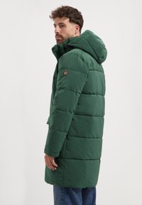 Man in een donkergroene gewatteerde pufferjas met een capuchon en horizontale stiksels, staande in profiel tegen een eenvoudige witte achtergrond.