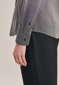 Grå långärmad skjorta med en subtil struktur, med en knäppning vid manschetten. I kombination med enfärgade svarta byxor. Sidovy av outfiten.