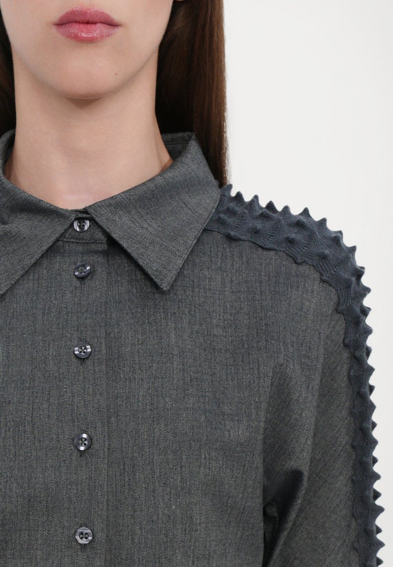 Grå skjorta med knappar och krage, som har texturerade volangdetaljer längs axlarna och mörka knappar på framsidan.
