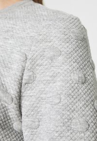 Tela textil gris con un patrón de diamantes texturizado y acentos circulares elevados, que exhibe un aspecto suave y cálido y un acabado liso.