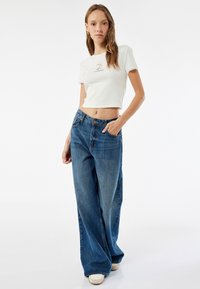 Hvid cropped t-shirt med bølgede kanter og blå grafik, parret med brede blå jeans. Modellen bærer hvide sneakers.