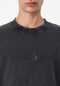 Dark gray cotton sweatshirt featuring a ribbed neckline and a raised black logo "HUGO" on the front. Smooth texture with a relaxed fit.