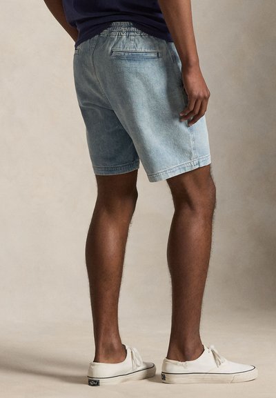 Άνδρας με ανοιχτό μπλε τζιν σόρτς, μπλε σκούρο πουκάμισο και λευκά χαμηλά αθλητικά παπούτσια, στέκεται σε ουδέτερο φόντο.